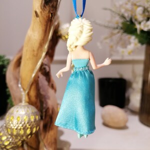 K&ouml;nnte beinhalten: Ein blauer und wei&szlig;er Plastik-Puppen-Anh&auml;nger einer Prinzessin mit blonden Haaren in einem Zopf, der an einem blauen Band h&auml;ngt.