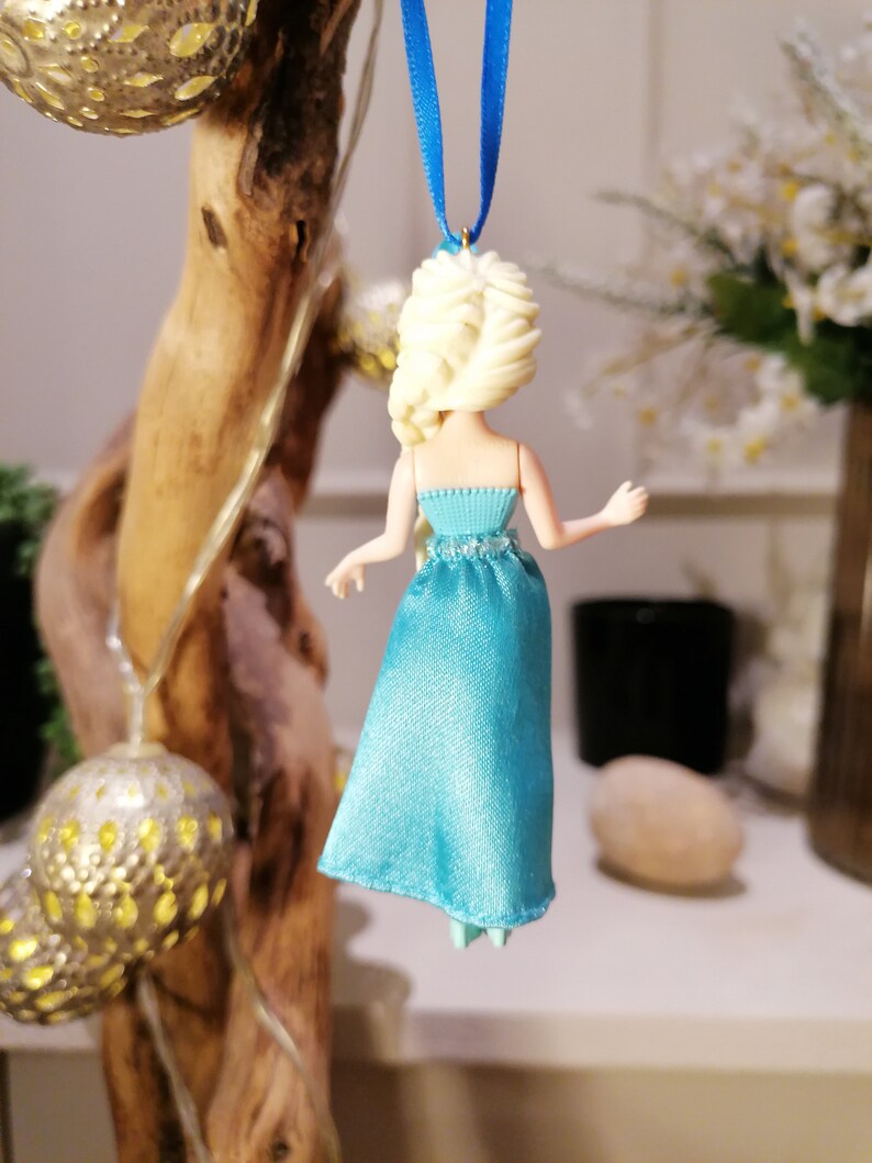 K&ouml;nnte beinhalten: Ein blauer und wei&szlig;er Plastik-Puppen-Anh&auml;nger einer Prinzessin mit blonden Haaren in einem Zopf, der an einem blauen Band h&auml;ngt.