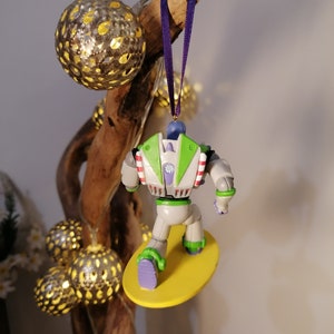 K&ouml;nnte beinhalten: Ein gr&uuml;ner und wei&szlig;er Buzz Lightyear Weihnachtsbaumschmuck mit einem lila Band, das an einem Ast h&auml;ngt. Buzz reitet auf einem gelben Surfbrett.