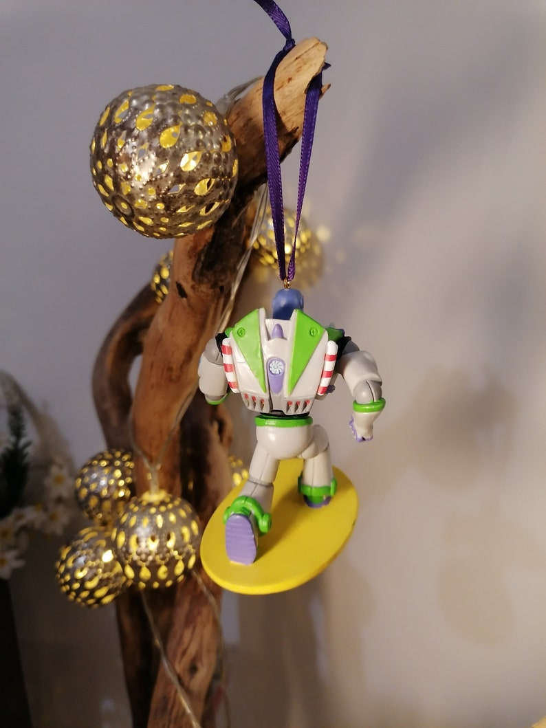 K&ouml;nnte beinhalten: Ein gr&uuml;ner und wei&szlig;er Buzz Lightyear Weihnachtsbaumschmuck mit einem lila Band, das an einem Ast h&auml;ngt. Buzz reitet auf einem gelben Surfbrett.