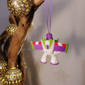 May include: A purple, white, and green plastic ornament of a cartoon space ranger with wings. The ornament is hanging from a purple ribbon.