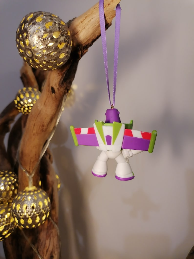 May include: A purple, white, and green plastic ornament of a cartoon space ranger with wings. The ornament is hanging from a purple ribbon.