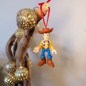 May include: A plastic toy figurine of Woody from Toy Story hanging from a branch with a red ribbon. Woody is wearing his signature cowboy hat, vest, and jeans.