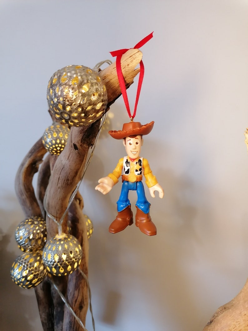 May include: A plastic toy figurine of Woody from Toy Story hanging from a branch with a red ribbon. Woody is wearing his signature cowboy hat, vest, and jeans.