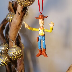 May include: A red string hangs from a wooden branch with a silver ornament. A plastic toy figurine of Woody from Toy Story is hanging from the string. Woody is wearing his signature cowboy hat, vest, and blue jeans. He is waving with his right hand and pointing with his left hand.