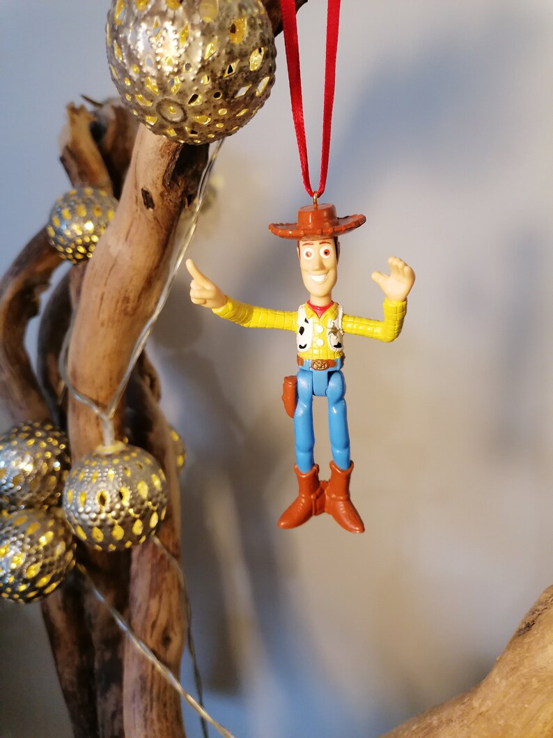May include: A red string hangs from a wooden branch with a silver ornament. A plastic toy figurine of Woody from Toy Story is hanging from the string. Woody is wearing his signature cowboy hat, vest, and blue jeans. He is waving with his right hand and pointing with his left hand.