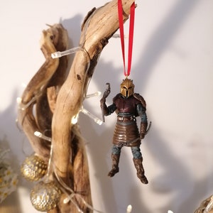 May include: A brown and gold action figure of a character wearing a helmet and armor, hanging from a red ribbon on a branch of driftwood. The figure is holding a weapon in one hand.