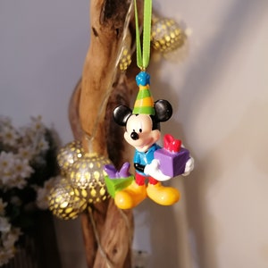 Peut inclure: Une petite figurine en plastique de Mickey Mouse portant un chapeau d'anniversaire et tenant une bo&icirc;te cadeau violette. La figurine est suspendue &agrave; un ruban vert.
