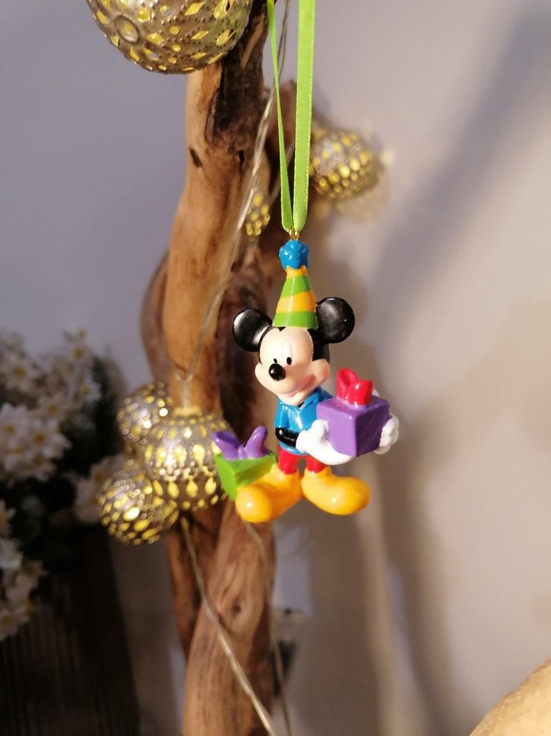 Peut inclure: Une petite figurine en plastique de Mickey Mouse portant un chapeau d'anniversaire et tenant une bo&icirc;te cadeau violette. La figurine est suspendue &agrave; un ruban vert.