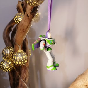 Peut inclure: Ornement de No&euml;l Buzz l'&Eacute;clair, avec une combinaison spatiale blanche, verte et violette, suspendu par un ruban violet. L'ornement est pr&egrave;s de lumi&egrave;res sph&eacute;riques d&eacute;coratives et d'une branche en bois naturel.