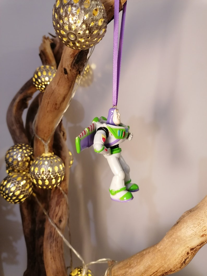Peut inclure: Ornement de No&euml;l Buzz l'&Eacute;clair, avec une combinaison spatiale blanche, verte et violette, suspendu par un ruban violet. L'ornement est pr&egrave;s de lumi&egrave;res sph&eacute;riques d&eacute;coratives et d'une branche en bois naturel.