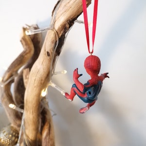 May include: A red and blue Spider-Man Christmas ornament hanging from a branch with white lights. The ornament is made of plastic and features a detailed design of Spider-Man in a climbing pose.