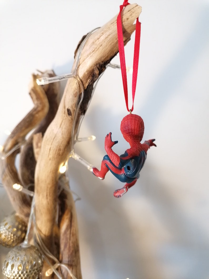 May include: A red and blue Spider-Man Christmas ornament hanging from a branch with white lights. The ornament is made of plastic and features a detailed design of Spider-Man in a climbing pose.