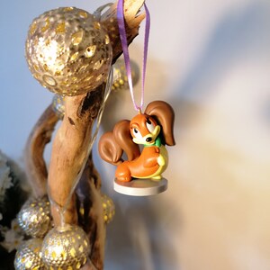 May include: A brown and orange squirrel figurine with a purple ribbon hanging from a branch with gold ornaments. The squirrel is wearing a green scarf and has a white base.