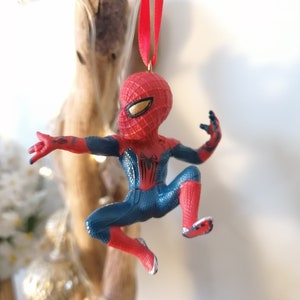 May include: Red and blue Spider-Man Christmas ornament hanging from a red ribbon. The ornament is a small figurine of Spider-Man in a web-slinging pose.