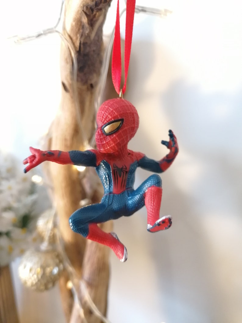 May include: Red and blue Spider-Man Christmas ornament hanging from a red ribbon. The ornament is a small figurine of Spider-Man in a web-slinging pose.