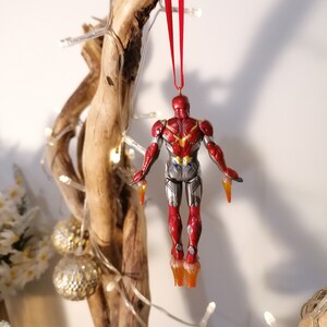 May include: Red and silver Iron Man Christmas ornament hanging from a red ribbon on a wooden branch. The ornament is in a flying pose with the arms outstretched and the legs slightly bent.