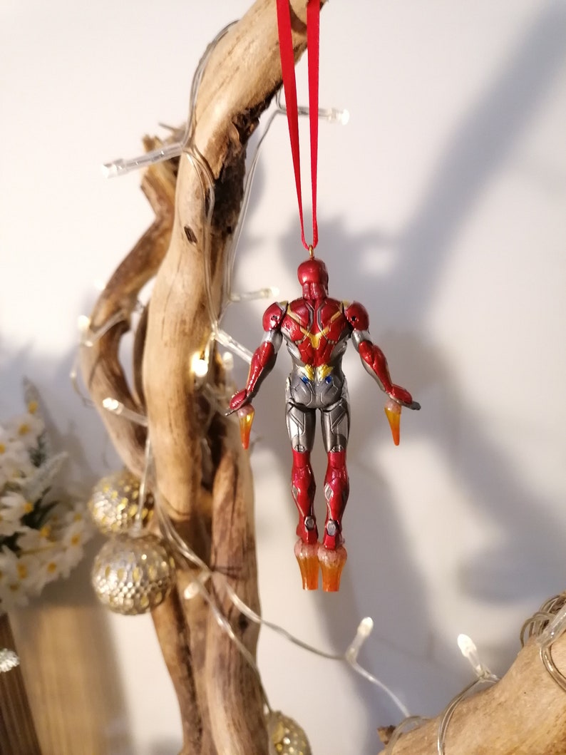 May include: Red and silver Iron Man Christmas ornament hanging from a red ribbon on a wooden branch. The ornament is in a flying pose with the arms outstretched and the legs slightly bent.