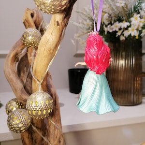 May include: A red-haired Disney princess doll ornament with a teal blue dress hanging from a branch with gold and white ornaments and a string of white lights.
