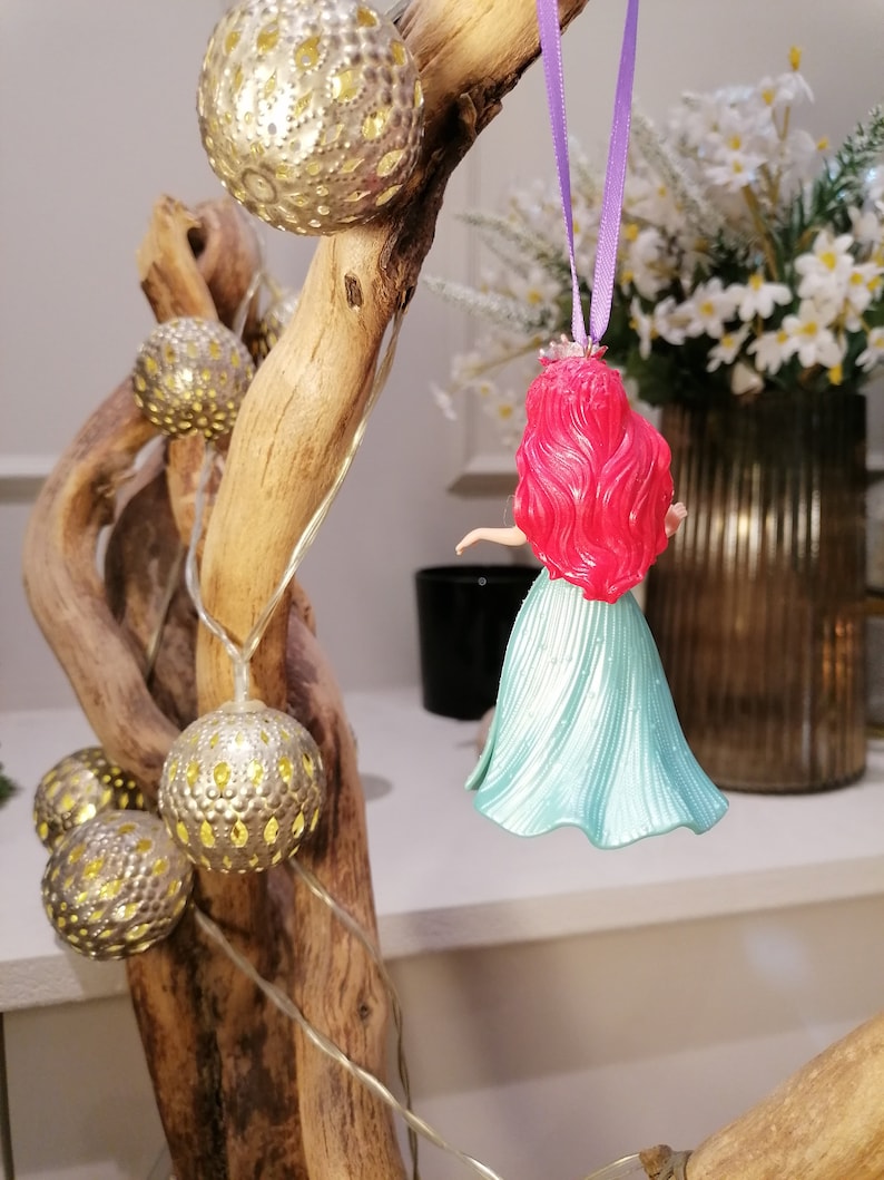 May include: A red-haired Disney princess doll ornament with a teal blue dress hanging from a branch with gold and white ornaments and a string of white lights.