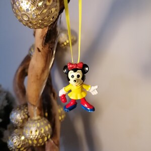 K&ouml;nnte beinhalten: Eine gelbe Schnur mit einer kleinen Plastikfigur von Minnie Maus, die daran h&auml;ngt. Minnie Maus tr&auml;gt ein gelbes Kleid mit roten Schuhen und einer roten Schleife. Die Schnur h&auml;ngt an einem St&uuml;ck Treibholz mit silbernen und goldenen Ornamenten.