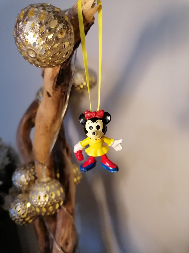 K&ouml;nnte beinhalten: Eine gelbe Schnur mit einer kleinen Plastikfigur von Minnie Maus, die daran h&auml;ngt. Minnie Maus tr&auml;gt ein gelbes Kleid mit roten Schuhen und einer roten Schleife. Die Schnur h&auml;ngt an einem St&uuml;ck Treibholz mit silbernen und goldenen Ornamenten.