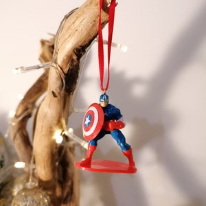 May include: A red and blue Captain America Christmas ornament with a red star on his chest and a red, white, and blue shield. The ornament is hanging from a red ribbon.