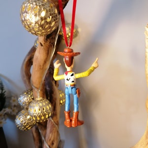 May include: A red string hangs from a piece of driftwood with a plastic toy figurine of Woody from Toy Story hanging from it. Woody is wearing his signature brown hat, yellow shirt, blue jeans, and brown boots. He is holding his right arm up in the air and his left arm is bent at the elbow.