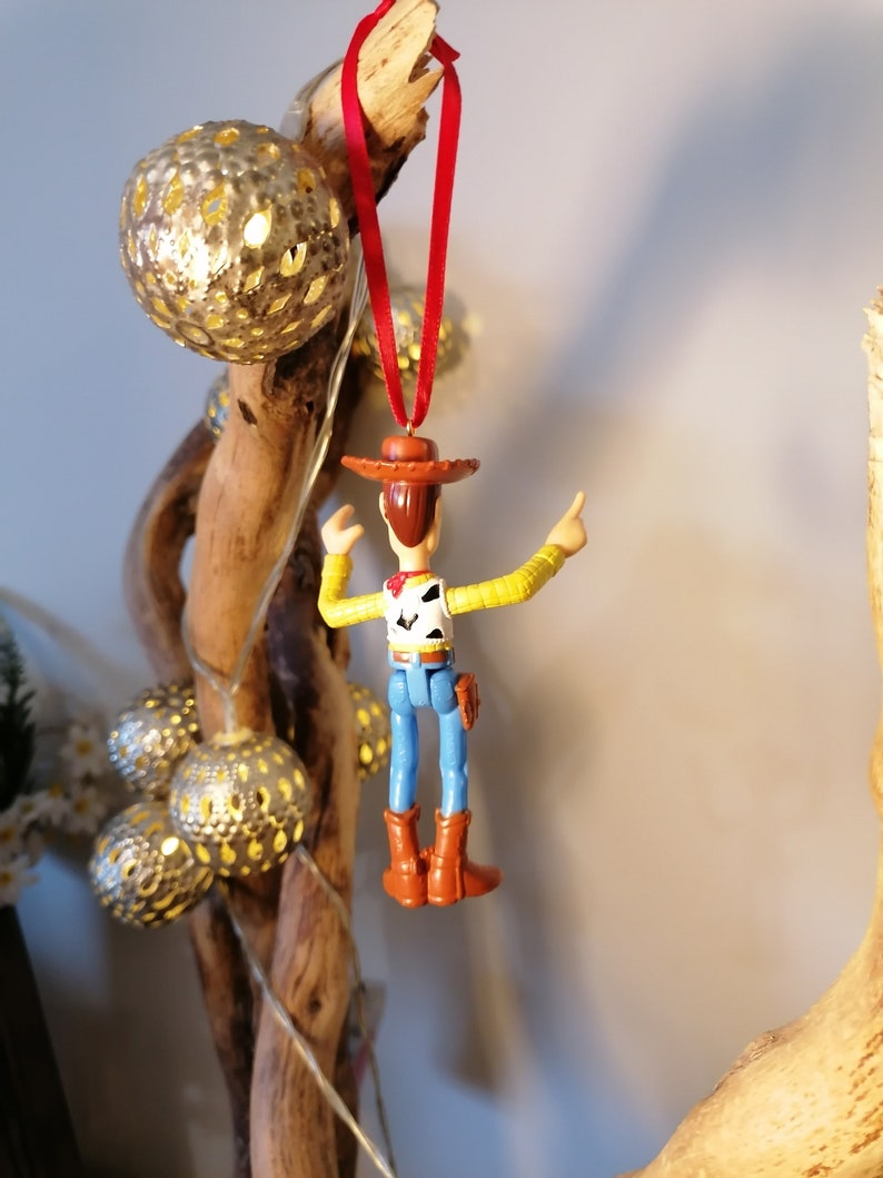 May include: A red string hangs from a piece of driftwood with a plastic toy figurine of Woody from Toy Story hanging from it. Woody is wearing his signature brown hat, yellow shirt, blue jeans, and brown boots. He is holding his right arm up in the air and his left arm is bent at the elbow.