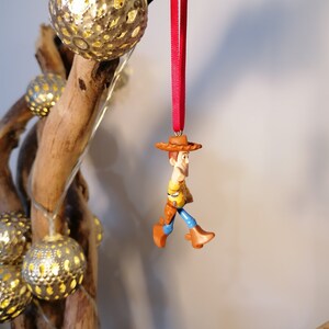 May include: A red string ornament featuring a plastic figurine of Woody from Toy Story. Woody is wearing his signature cowboy hat, vest, and jeans. He is holding his lasso and has a friendly expression on his face.