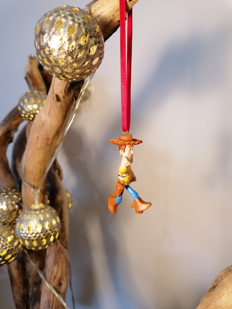 May include: A red string ornament featuring a plastic figurine of Woody from Toy Story. Woody is wearing his signature cowboy hat, vest, and jeans. He is holding his lasso and has a friendly expression on his face.