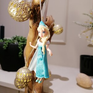 May include: A blue and white plastic doll with blonde hair wearing a blue dress and a crown. The doll is hanging from a branch with a white string.