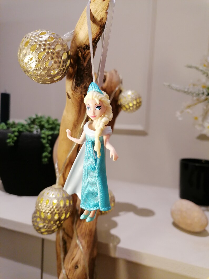 May include: A blue and white plastic doll with blonde hair wearing a blue dress and a crown. The doll is hanging from a branch with a white string.