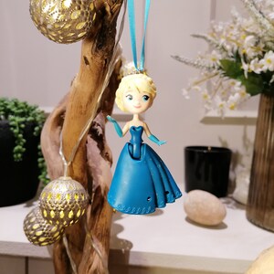 Peut inclure: Une poup&eacute;e en plastique bleue repr&eacute;sentant une princesse avec une robe bleue et des cheveux blonds, suspendue &agrave; un ruban bleu. La poup&eacute;e est suspendue &agrave; une branche de bois flott&eacute; avec des boules d&eacute;coratives argent&eacute;es et dor&eacute;es.