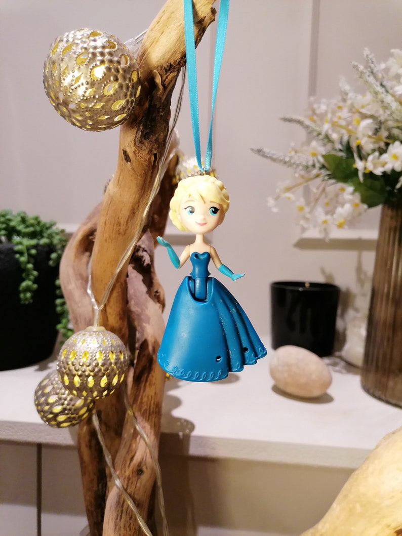Peut inclure: Une poup&eacute;e en plastique bleue repr&eacute;sentant une princesse avec une robe bleue et des cheveux blonds, suspendue &agrave; un ruban bleu. La poup&eacute;e est suspendue &agrave; une branche de bois flott&eacute; avec des boules d&eacute;coratives argent&eacute;es et dor&eacute;es.
