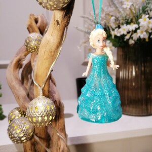 May include: A blue glitter ornament featuring a princess doll with blonde hair and a blue dress. The ornament is hanging from a blue ribbon on a wooden branch with string lights.