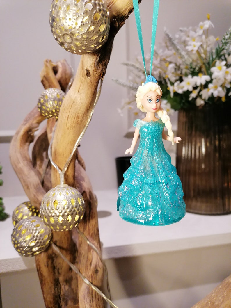 May include: A blue glitter ornament featuring a princess doll with blonde hair and a blue dress. The ornament is hanging from a blue ribbon on a wooden branch with string lights.