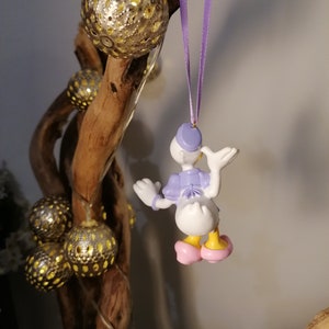 May include: A purple and white figurine of a cartoon duck wearing a purple hat and pink shoes. The figurine is hanging from a purple ribbon on a wooden branch with gold ornaments.