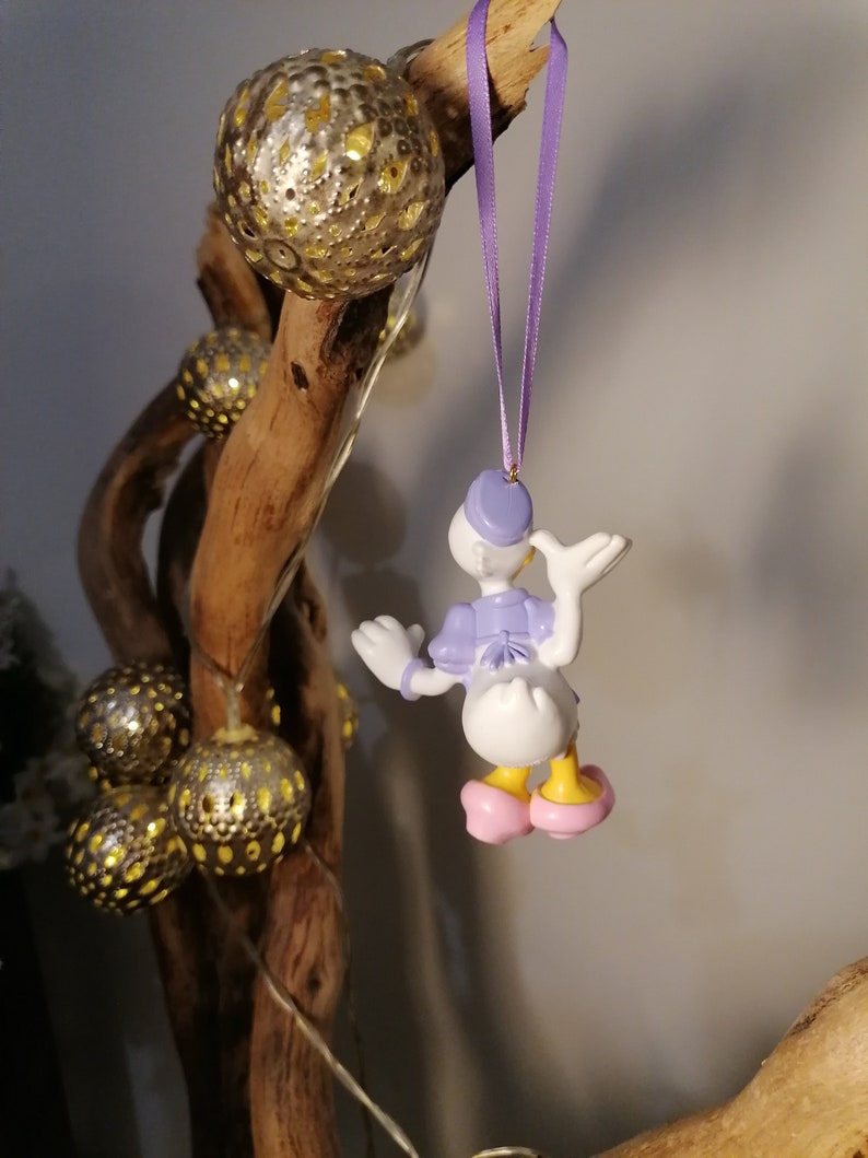 May include: A purple and white figurine of a cartoon duck wearing a purple hat and pink shoes. The figurine is hanging from a purple ribbon on a wooden branch with gold ornaments.