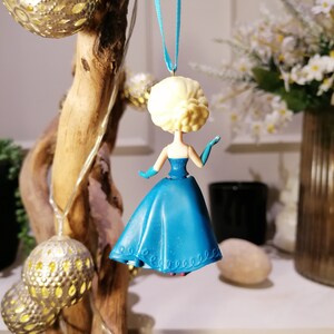 Peut inclure: Une d&eacute;coration de No&euml;l bleue et blanche repr&eacute;sentant une figurine de princesse v&ecirc;tue d'une robe bleue et d'une couronne blanche. La d&eacute;coration est suspendue &agrave; un ruban bleu.