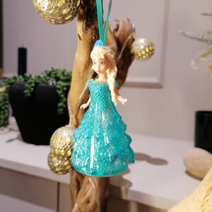 May include: A blue and white plastic doll with blonde hair wearing a sparkly blue dress. The doll is hanging from a branch with a blue ribbon.