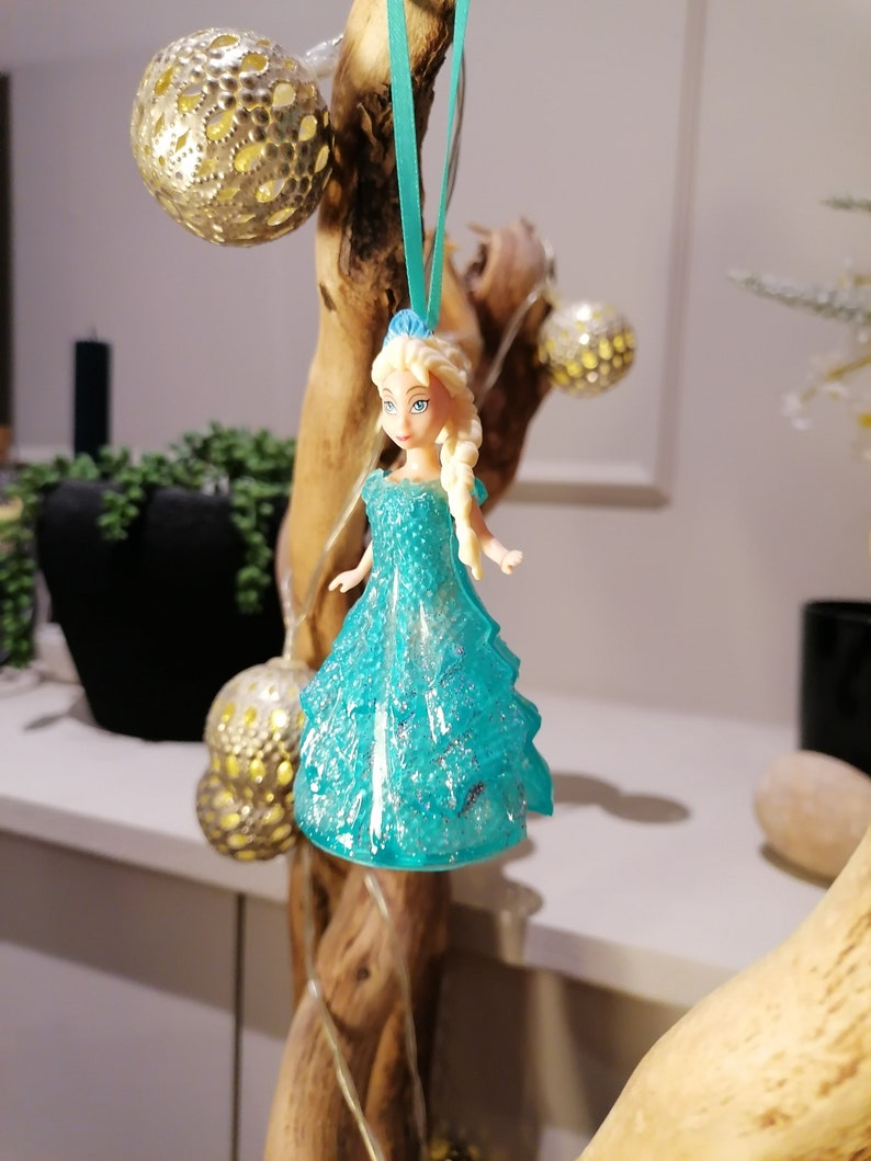 May include: A blue and white plastic doll with blonde hair wearing a sparkly blue dress. The doll is hanging from a branch with a blue ribbon.