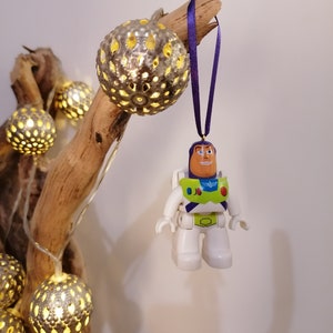 May include: A white and green Buzz Lightyear toy ornament hanging from a branch with a purple ribbon. The ornament is made of plastic and has a detailed design. The branch is decorated with string lights and silver metallic balls.