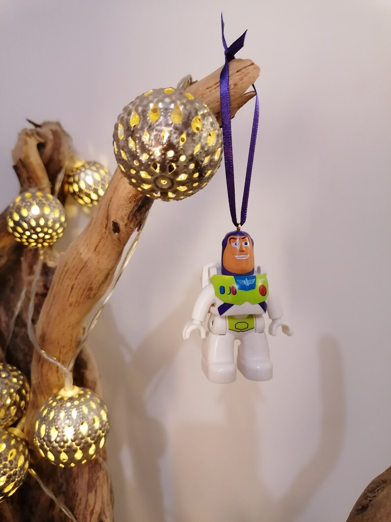 May include: A white and green Buzz Lightyear toy ornament hanging from a branch with a purple ribbon. The ornament is made of plastic and has a detailed design. The branch is decorated with string lights and silver metallic balls.