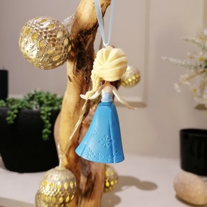 May include: A blue and white plastic ornament featuring a small doll with blonde hair and a blue dress. The doll is hanging from a blue ribbon on a wooden branch.