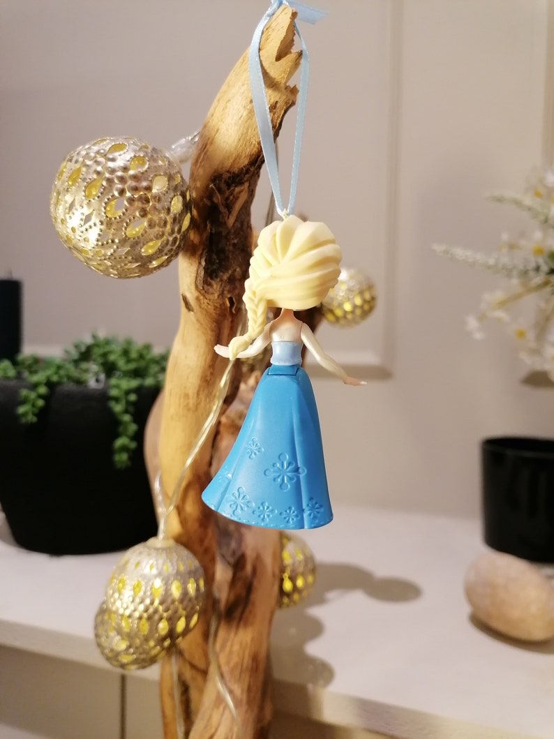 May include: A blue and white plastic ornament featuring a small doll with blonde hair and a blue dress. The doll is hanging from a blue ribbon on a wooden branch.