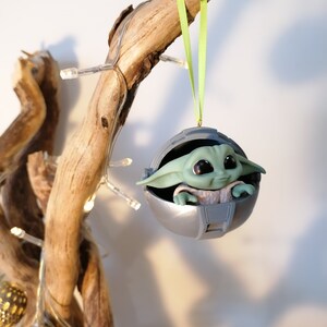 May include: A green and gray Christmas ornament featuring a character from the Star Wars franchise, The Mandalorian. The character is sitting in a round, silver pod with a green ribbon attached.