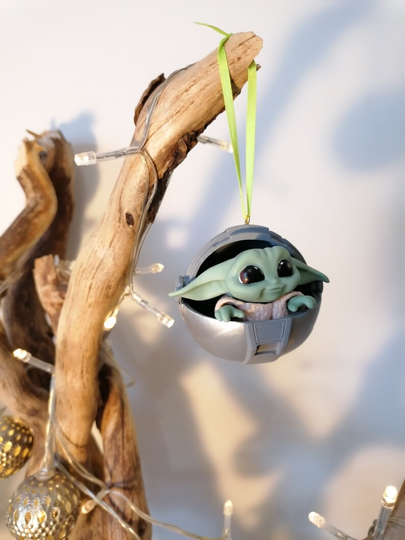May include: A green and gray Christmas ornament featuring a character from the Star Wars franchise, The Mandalorian. The character is sitting in a round, silver pod with a green ribbon attached.