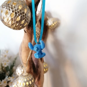 May include: A blue Stitch character charm hanging from a turquoise ribbon. The charm is attached to a wooden branch with string lights wrapped around it.