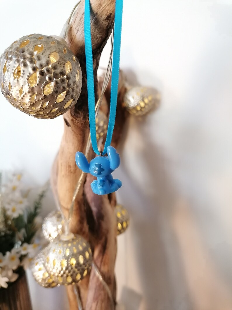 May include: A blue Stitch character charm hanging from a turquoise ribbon. The charm is attached to a wooden branch with string lights wrapped around it.
